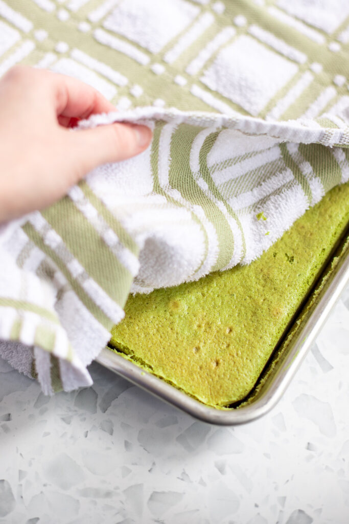 Steaming matcha cake with a tea towel on top, hand lifting to show the sponge.