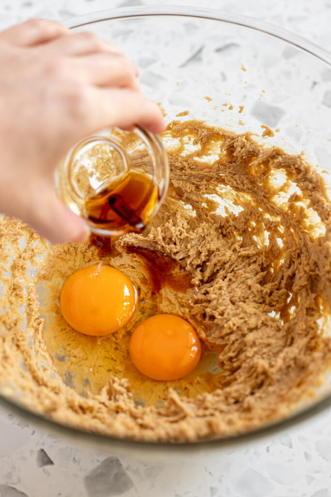 Hand tipping vanilla extract into bowl containing butter, sugar, and eggs.