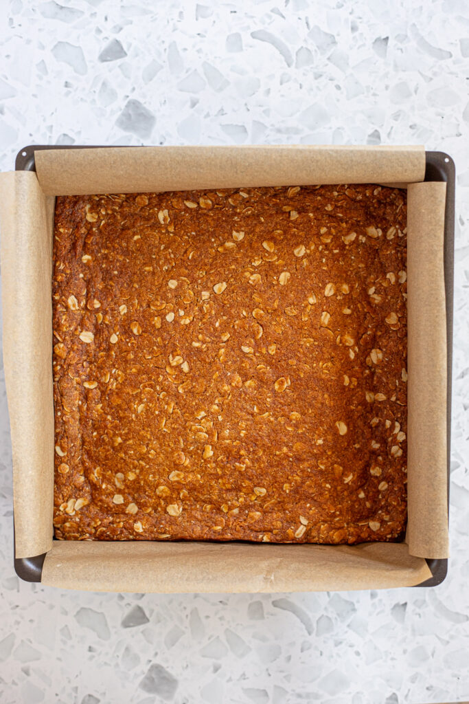 Top-down view of freshly baked Anzac slice in parchment-lined tin, golden brown with oats visible