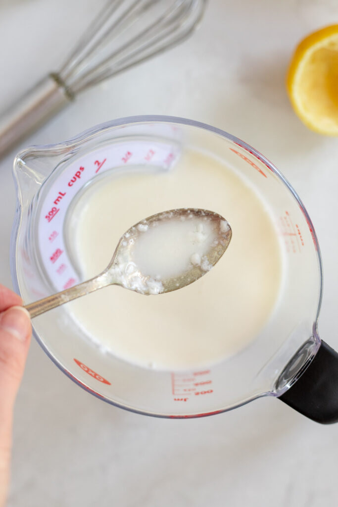 Hand holding a spoon of curdled DIY buttermilk over a measuring jug, with a whisk and squeezed lemon half nearby