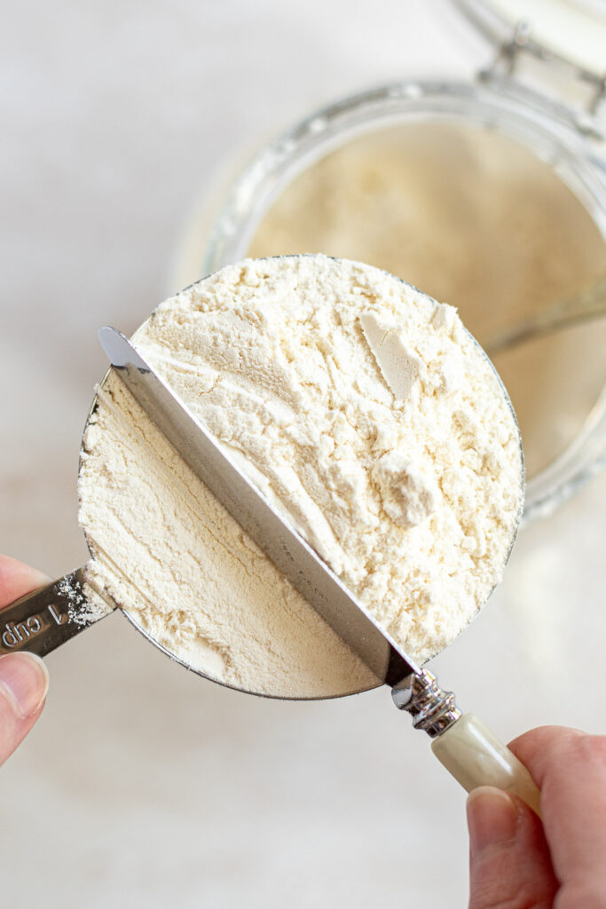 Using a knife to level flour in a measuring cup, scraping the excess back into the container.