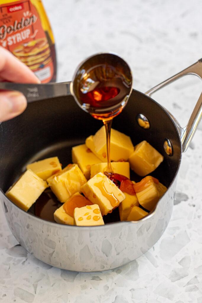 Using a tablespoon to drizzle golden syrup into a saucepan containing cubes of butter