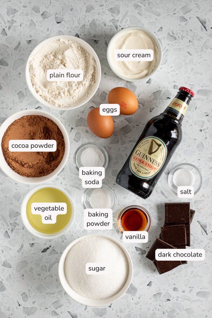 Labelled flatlay of chocolate Guinness cake ingredients, including a bottle of Guinness, dark chocolate, and cocoa powder