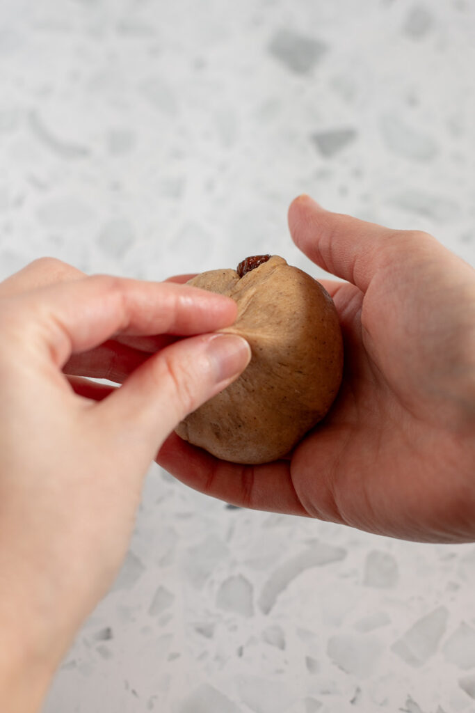 Hands shaping hot cross bun dough into smooth balls, pinching the bottom to seal like a money bag