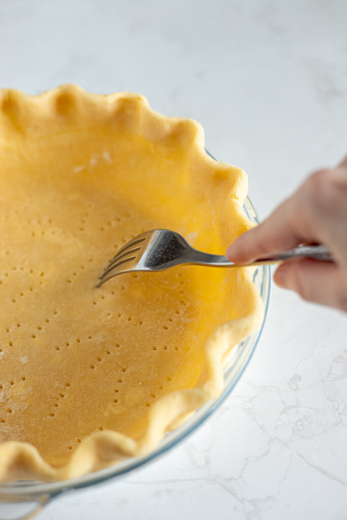 Close-up of a hand pricking holes in an unbaked pie crust with a fork before blind baking.