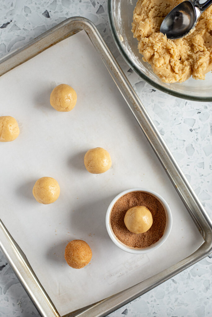 Top-down view of cookie dough balls being rolled in cinnamon sugar, with bowl of dough and scoop behind