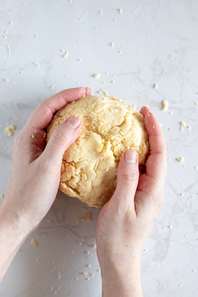 Hands bringing crumbly pie dough together into a rough ball.