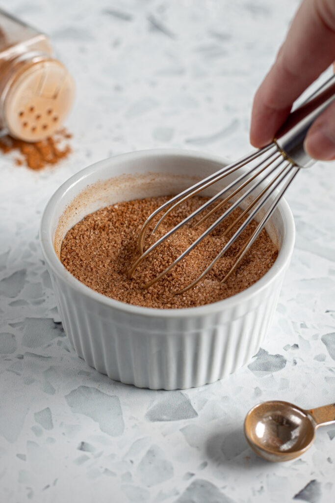 Hand whisking cinnamon sugar in a small ramekin, with a teaspoon and a shaker of ground cinnamon nearby