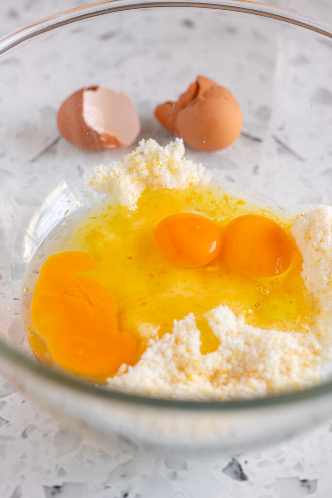 Three eggs cracked into orange-infused sugar with eggshells in the background.