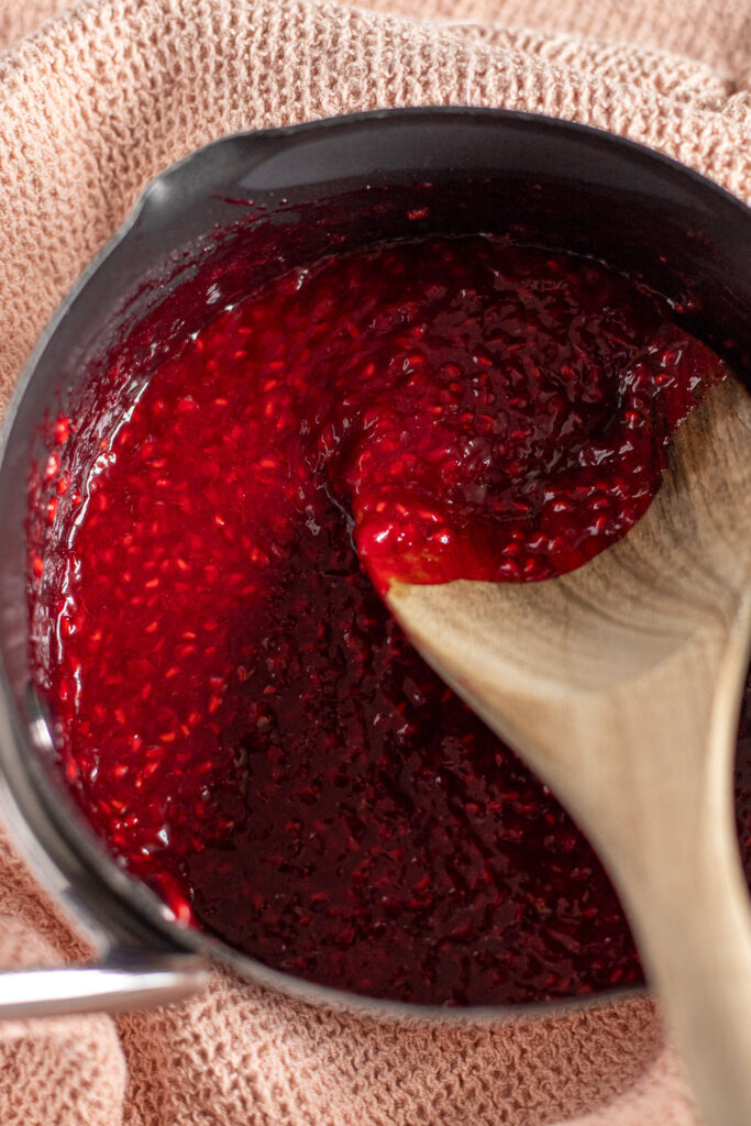 Top-down view of glossy, ruby-red raspberry pie filling in a saucepan, with visible seeds and a wooden spoon resting inside.