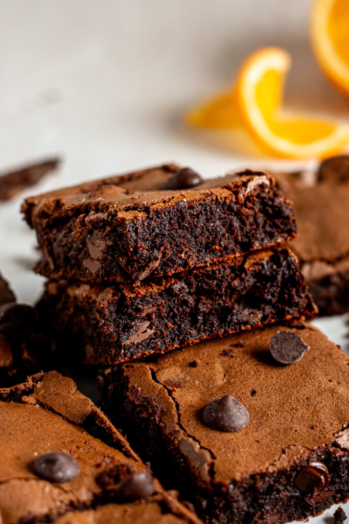 Two flourless chocolate orange brownies stacked at an angle to reveal their fudgy interior, with a twisted orange slice and scattered dark chocolate chips nearby.