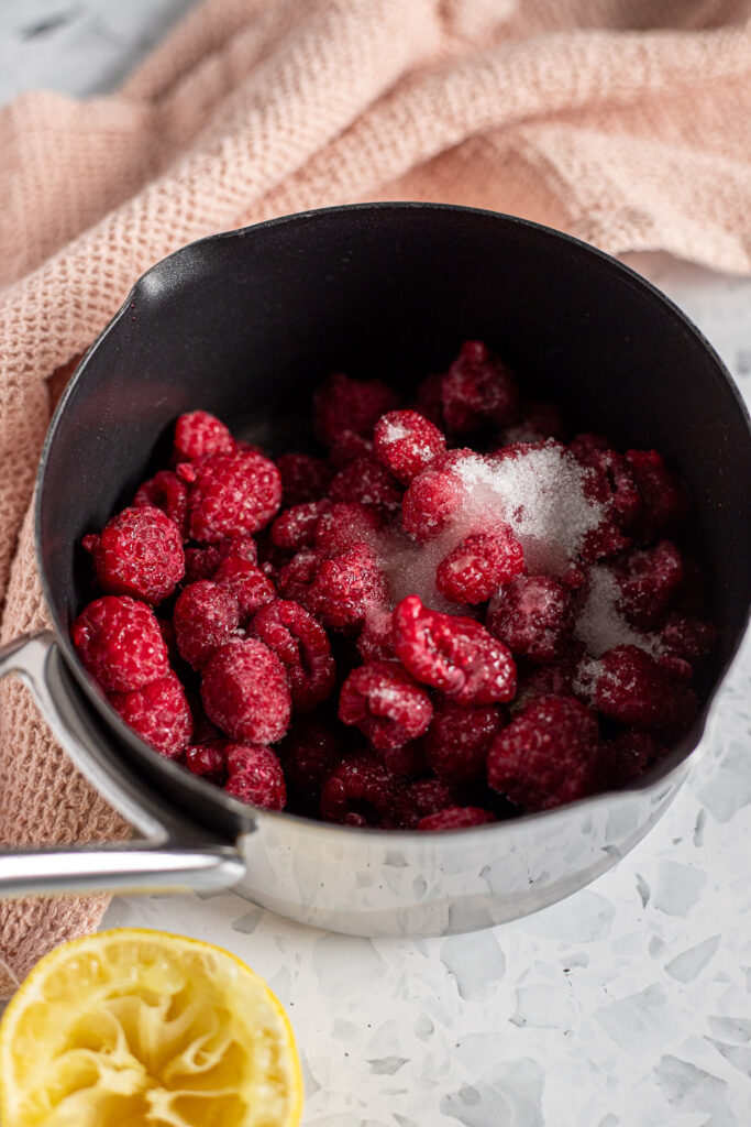 Fresh raspberries, sugar, and lemon juice in a small saucepan, styled with pink linens and a squeezed lemon half.
