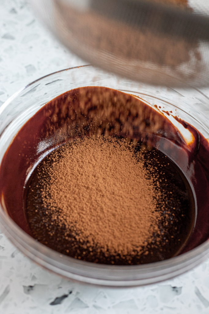 Sifting cocoa powder into a glass bowl of melted chocolate and butter.