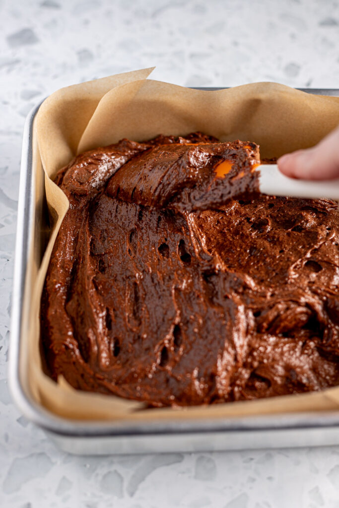 Spreading thick chocolate orange brownie batter evenly into a parchment-lined square metal tin.