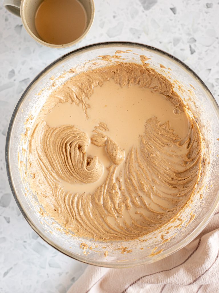 Chai-infused milk being gradually added to the cake batter, alternating with the dry ingredients, with a mug beside the bowl.