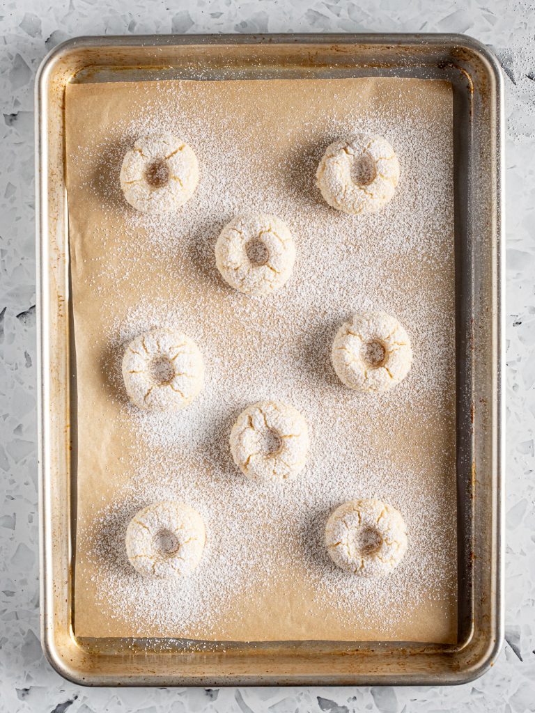 Baked amaretti thumbprint cookies with cracked, puffed tops, dusted with icing sugar on a baking tray.