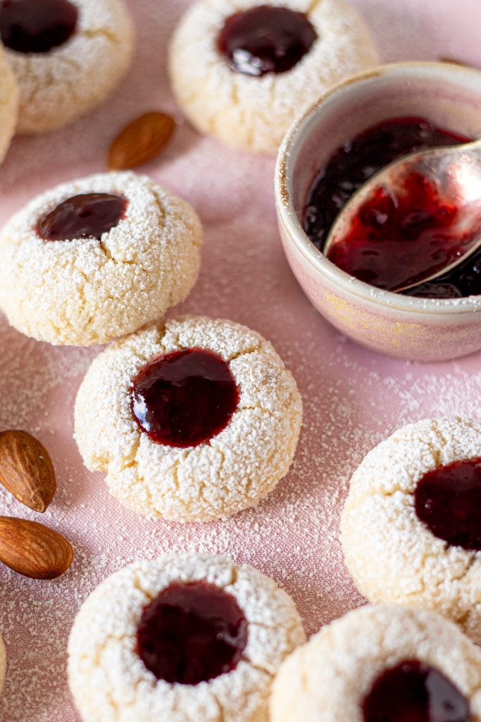 Cherry amaretti jam drops arranged on pink linen with whole almonds, icing sugar and a small pink ramekin of cherry jam with a spoon.