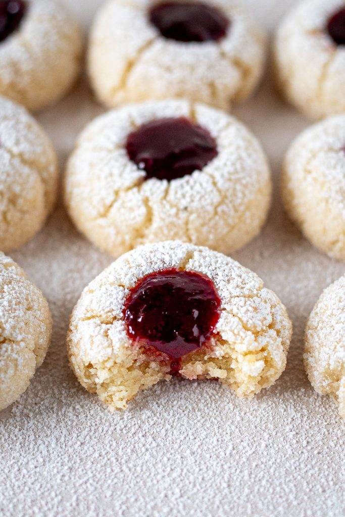 Cherry amaretti jam drops arranged in neat rows on a light beige background, with a bite taken out of the centre cookie revealing its interior and cherry jam filling.