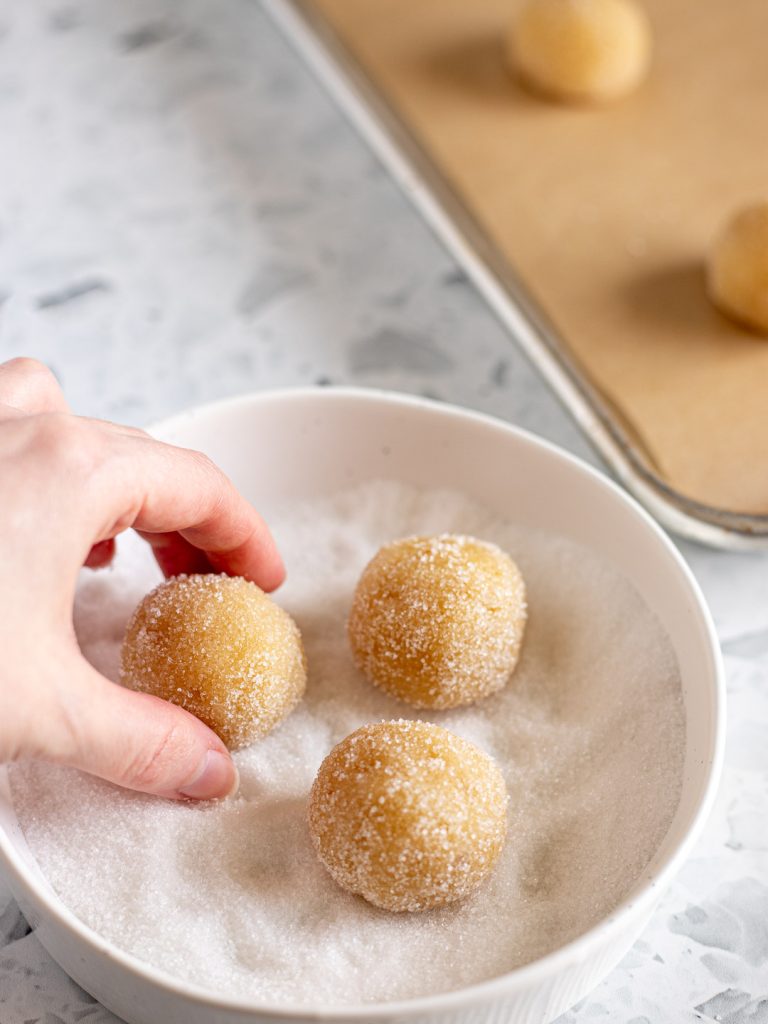 Amaretti dough balls being rolled in a light coating of sugar before being placed on a lined baking tray.