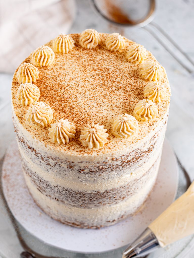 Vanilla chai cake on a turntable with a piped buttercream border around the top edge, dusted with cinnamon, with a piping bag and sieve nearby.