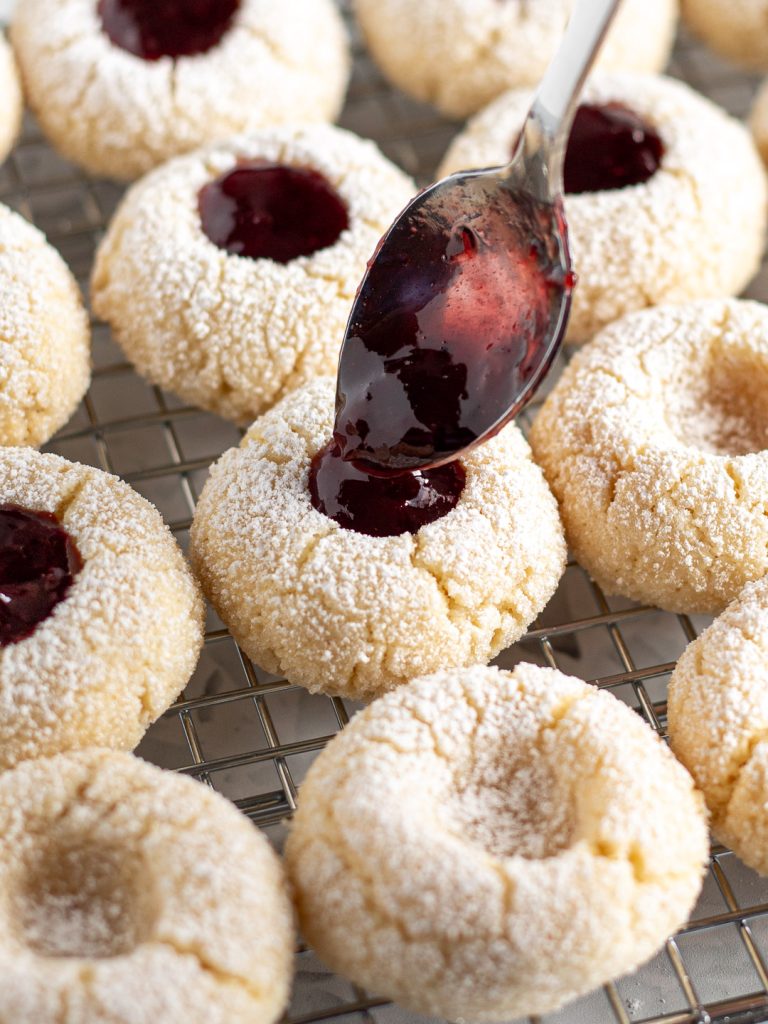 Cherry jam being spooned into the indents of baked amaretti thumbprint cookies resting on a cooling rack.