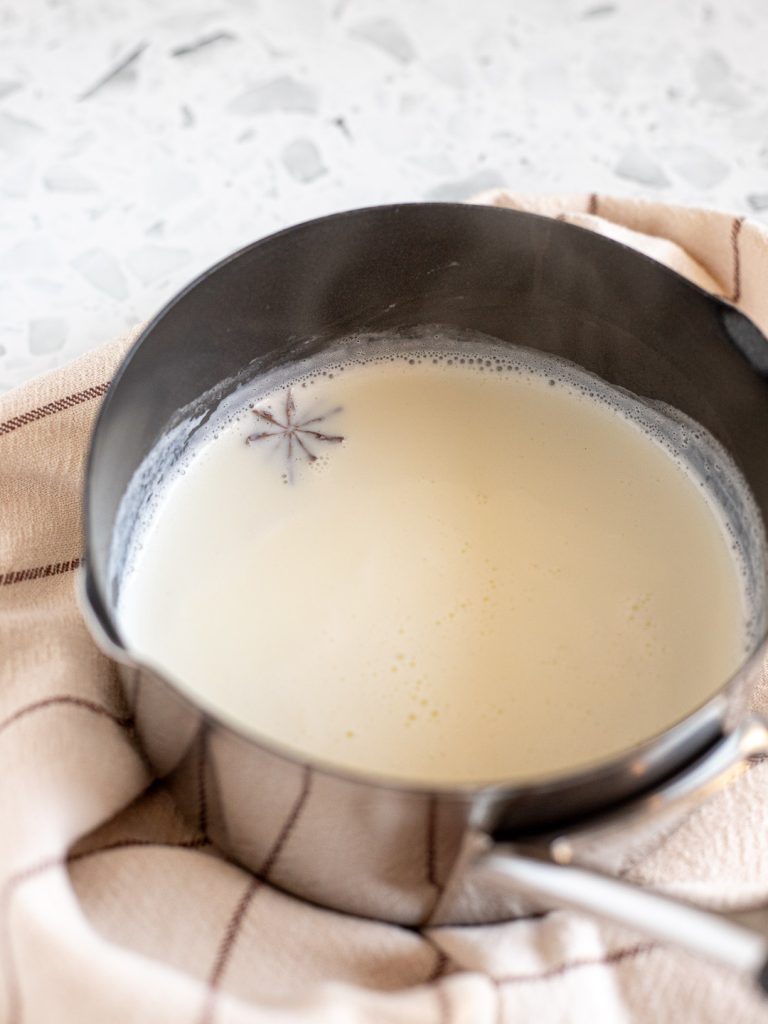 Warm, foamy milk with star anise in a small saucepan on a beige striped tea towel.