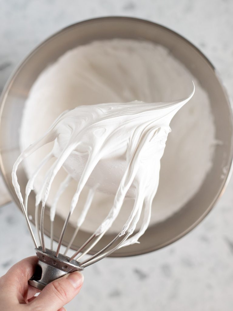 Hand holding a whisk attachment with stiff peaks of meringue above a stainless steel mixer bowl.