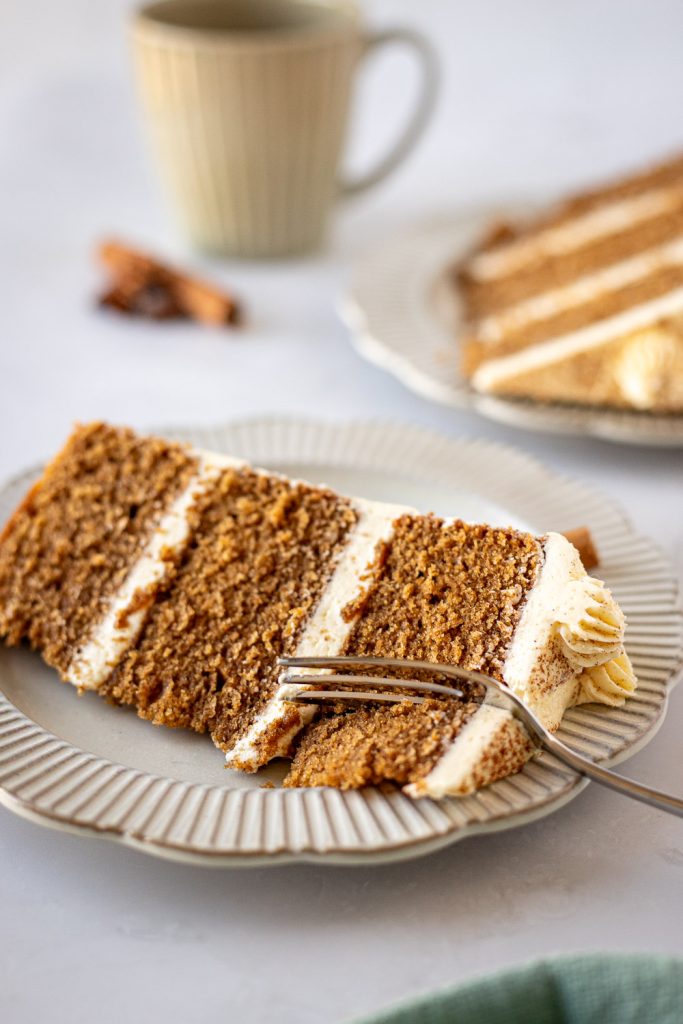Three-layer vanilla chai cake slice on a scalloped plate, with a fork cutting into the soft sponge and another slice and a mug in the background.