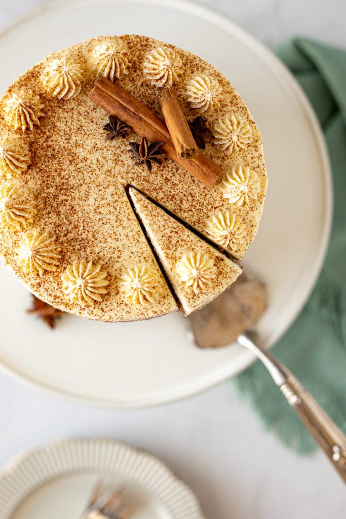 Top-down view of vanilla chai cake with piped borders, a dusting of cinnamon, star anise and cinnamon sticks, as a slice is lifted with a cake server. A teal linen surrounds the cake stand.